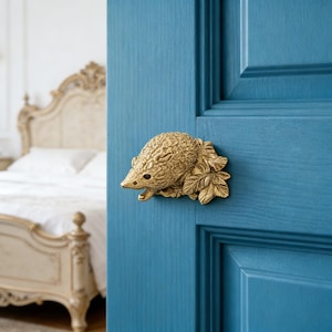 Könnte beinhalten: Ein goldfarbener Türknauf in Form eines Igels, der an einer blauen Tür befestigt ist. Der Türknauf ist eine detaillierte Skulptur eines Igels mit Blättern. Im Hintergrund ist ein Bett mit einer weißen Bettdecke zu sehen.