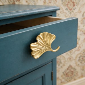 Puede incluir: Un mueble azul verdoso con un cajón ligeramente abierto, que muestra un tirador dorado en forma de hoja de ginkgo. El mueble tiene un acabado envejecido y el interior del cajón es de color madera clara. El tirador es el punto focal.