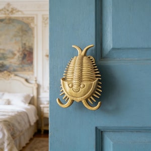 May include: A gold-coloured trilobite-shaped door knocker fixed to a light blue door. The knocker features a detailed, textured surface and a smiling face design. The door is part of a room with a bed and ornate decor.
