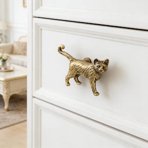 Op de afbeelding: Een messing katvormige trekker is op een witte lade gemonteerd. De kat wordt afgebeeld in een loopstand met gekrulde staart. De trekker voegt een decoratief tintje toe aan meubels.
