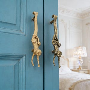 May include: Two brass monkey-shaped door handles fixed to a teal blue door. The handles are detailed and decorative. The door is open, revealing a bedroom with a bed and a lamp.