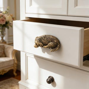 May include: A white dresser drawer with a bronze-colored bear-shaped handle. The drawer is open, revealing the wooden interior. A second, closed drawer with a dark knob is visible below.