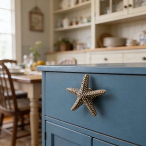Peut inclure: Un meuble bleu avec une poignée en forme d'étoile de mer. La poignée est de couleur marron clair avec de petits détails texturés. Le meuble est dans une cuisine avec une table et des chaises en bois en arrière-plan.