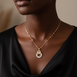 May include: A gold-colored necklace with a pendant featuring two interlocking circles encrusted with small, clear gemstones. The necklace is worn on a person with dark skin, and the background is a soft, neutral tone.