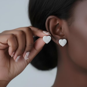 May include: Heart-shaped stud earrings, each encrusted with small, sparkling stones. One earring is held up to an ear, the other is in place. The earrings are set against a plain background, showcasing the jewellery's design.