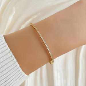 May include: A gold-colored bracelet with a row of small, clear gemstones. The bracelet is worn on a wrist, and the background is a soft, white fabric. The bracelet appears to be approximately 7 inches in circumference.