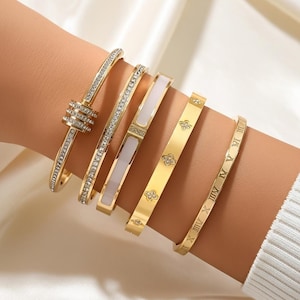 May include: A collection of gold-toned bangles, each with unique designs. Some are adorned with clear crystals, while others feature Roman numerals, floral patterns, or white enamel accents. The bangles are displayed on a wrist.