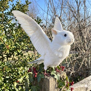 Pu&ograve; includere: Una scultura di coniglio bianco alato con le ali spiegate, appollaiata su un palo di legno ornato di edera e bacche rosse. La scultura ha un corpo soffice, occhi neri e piume dettagliate sulle ali.