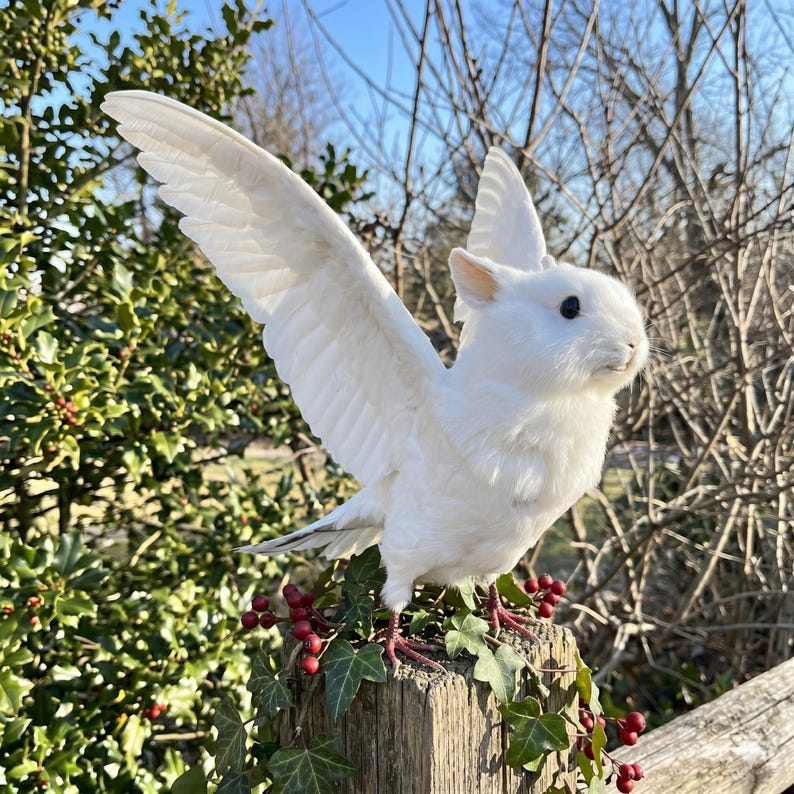 Pu&ograve; includere: Una scultura di coniglio bianco alato con le ali spiegate, appollaiata su un palo di legno ornato di edera e bacche rosse. La scultura ha un corpo soffice, occhi neri e piume dettagliate sulle ali.