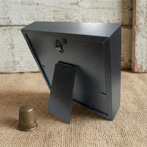 May include: A black picture frame with a built-in easel back, angled to show the back of the frame. A small, brass thimble sits in front of the frame. The frame is on a burlap surface.