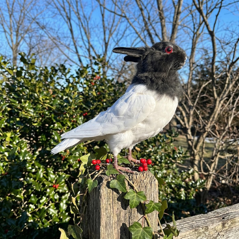 Pu&ograve; includere: Una scultura unica di un uccello con la testa di un coniglio, appollaiata su un palo di legno. Il corpo dell'uccello &egrave; bianco, con testa e collo neri. Bacche rosse e foglie verdi adornano il palo, sullo sfondo di alberi e cielo azzurro.