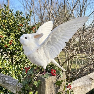 Pu&ograve; includere: Una scultura di coniglio bianco con grandi ali piumate, appollaiata su un palo di legno adornato con edera e bacche rosse. Il coniglio ha orecchie rosa e occhi neri. Le ali sono spiegate, su uno sfondo di vegetazione e cielo blu.