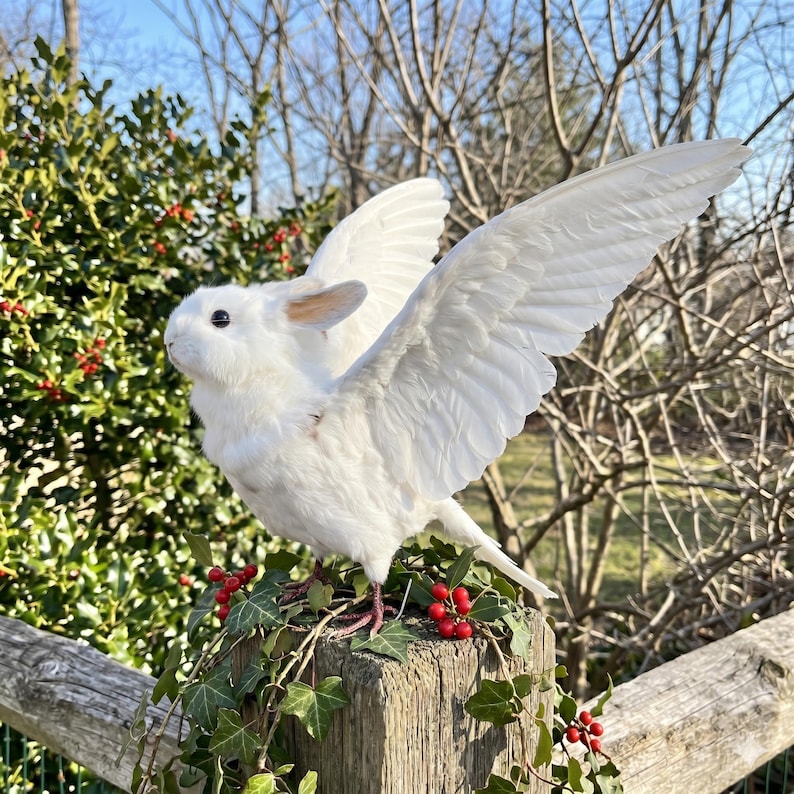 Pu&ograve; includere: Una scultura di coniglio bianco con grandi ali piumate, appollaiata su un palo di legno adornato con edera e bacche rosse. Il coniglio ha orecchie rosa e occhi neri. Le ali sono spiegate, su uno sfondo di vegetazione e cielo blu.