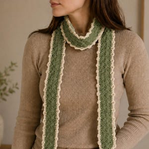 May include: A long, narrow crochet scarf featuring a textured sage green center panel with a delicate, scalloped cream-colored border along all edges. The scarf is draped around the neck, hanging down in two even vertical strips over a beige ribbed sweater.