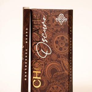 Könnte beinhalten: Dunkelbraune Schokoladenverpackung mit goldfarbener Aufschrift "CHOCOLATE Oscuro 70%" und "TRAPICHE cacao". Die Verpackung hat ein Zahnrad-Design und die Worte "CACAO VENEZOLANO" an der Seite.