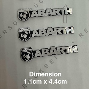 Może przedstawiać: Cztery srebrno-czarne emblematy samochodowe Abarth. Każdy emblemat zawiera logo Abarth i napis "ABARTH". Wymiary to 1,1 cm x 4,4 cm.