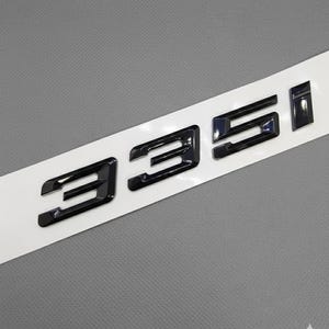 Può includere: Emblema 335i nero su una striscia bianca. L'emblema è lucido e sembra essere in plastica o metallo. Lo sfondo è di colore grigio uniforme.