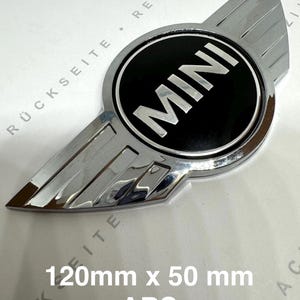 Może przedstawiać: Chromowany emblemat MINI z czarnym okrągłym środkiem. Słowo "MINI" jest w białych, pogrubionych literach. Emblemat ma rozszerzenia w kształcie skrzydeł. Tekst "120mm x 50 mm ABS" znajduje się poniżej.