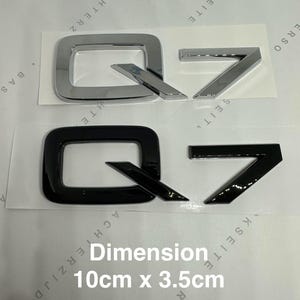 Op de afbeelding: Twee Q7-embleembadges, één chroom en één zwart, worden weergegeven op een wit oppervlak. De tekst "Dimension 10cm x 3.5cm" is zichtbaar. De badges meten 10 cm bij 3,5 cm.