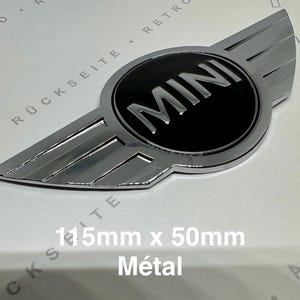 May include: A chrome and black MINI emblem with wing design. The emblem is 115mm wide and 50mm tall. The word "MINI" is in silver lettering on a black background. The word "Metal" is written below the emblem.