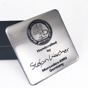 May include: A square, brushed metal plaque with the AMG logo and the text "Handcrafted by Stefan Karcher Mercedes-AMG Germany". The logo is a circular emblem with a tree and the words "Affalterbach" and "AMG".