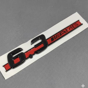 Peut inclure: Emblème AMG 6.3 noir et rouge sur un support blanc. L'emblème présente le texte "6.3" en noir et "AMG" en rouge. Le fond est une surface gris clair.
