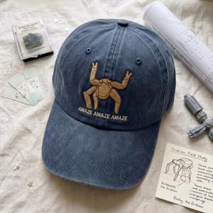 May include: A blue denim baseball cap featuring an embroidered tan creature with multiple limbs centered on the front panel. Below the creature, the text "AMAZE AMAZE AMAZE" is stitched in a matching tan thread. The cap has a curved brim.
