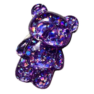 May include: A purple glitter teddy bear figurine. The bear is made of a translucent material and filled with iridescent glitter. The bear has a rounded head, small ears, and a simple body shape. The glitter reflects various colors.