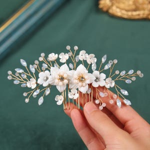 May include: A gold-toned hair comb adorned with white fabric flowers, clear crystals, and small pearl-like beads. The floral arrangement includes larger, multi-petaled flowers and smaller blossoms, creating a delicate, elegant design. The comb is held against a dark green background.