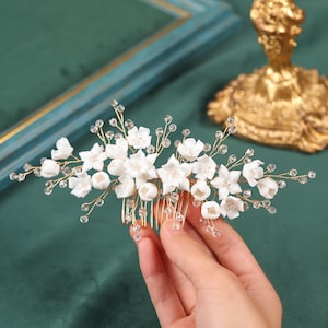 Könnte beinhalten: Ein goldener Haarkamm mit weißen Porzellanblüten und funkelnden Kristallakzenten. Das florale Design ist auf einem goldenen Kamm angeordnet, ideal für Hochzeiten oder besondere Anlässe. Länge ca. 10 cm.