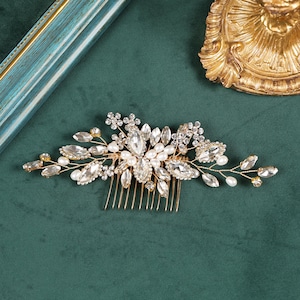 Könnte beinhalten: Ein goldfarbener Haarkamm mit einer dekorativen Anordnung aus klaren, marquise-geschliffenen Kristallen, kleinen runden Strasssteinen und weißen Kunstperlen. Das Design ist auf einem dünnen Golddrahtrahmen mit einer Metallkammbasis befestigt.