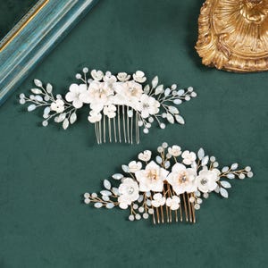 Op de afbeelding: Twee haarkammen met witte bloemaccenten, heldere kristallen in het midden en fijne draadtakjes versierd met kleine iriserende kralen. De bovenste kam heeft een zilverkleurige basis, terwijl de onderste kam een goudkleurige metalen basis heeft.