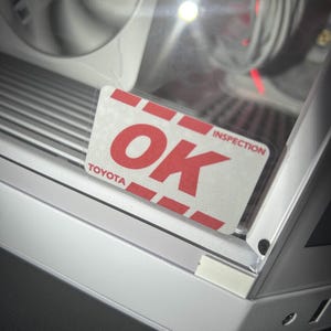 May include: A close-up of a white and silver computer case with a clear side panel. A red and white "OK INSPECTION TOYOTA" sticker is affixed to the panel. The case has a fan and internal wiring visible.