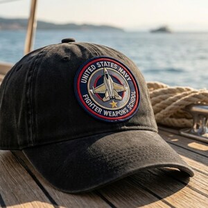 May include: A charcoal grey baseball cap featuring a circular embroidered patch on the front. The patch displays a fighter jet graphic, a gold star, and the text United States Navy Fighter Weapons School in a blue, red, and gold colour scheme.