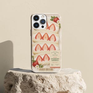 May include: A smartphone case featuring a graphic print of three Japanese strawberry sandwiches with white cream and sliced red strawberries. A rectangular label reads SOLARA MARKET FRESH PICKS JAPANESE STRAWBERRY SANDWICH.