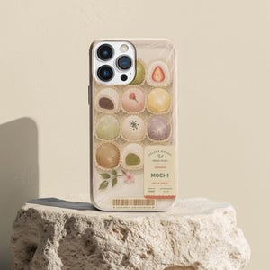 May include: A phone case featuring a grid of various mochi treats in paper cups, including a strawberry-filled piece. The design includes a label with the text SOLANA MARKET, FRESH PICKS, JAPANESE MOCHI, SOFT & CHEWY, and a barcode at the bottom.