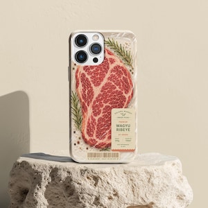 May include: A smartphone case featuring a graphic print of a raw Wagyu ribeye steak with rosemary sprigs and peppercorns. The design includes a label reading Solara Market, Premium Wagyu Ribeye, A5 Grade, 300g, and a barcode at the bottom edge.