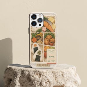 May include: A smartphone case featuring a detailed illustration of a Japanese bento box with rice balls, salmon, fried chicken, and vegetables. A label on the case reads SOLARA MARKET FRESH PICKS JAPANESE BENTO BALANCED & DELICIOUS TODAY.