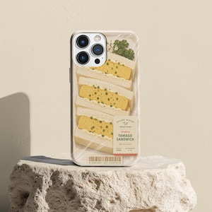 May include: A smartphone case featuring a graphic print of a Japanese tamago sandwich wrapped in plastic. The design includes a label with the text SOLARA MARKET, FRESH PICKS, JAPANESE TAMAGO SANDWICH, SOFT & FLUFFY, and a barcode.