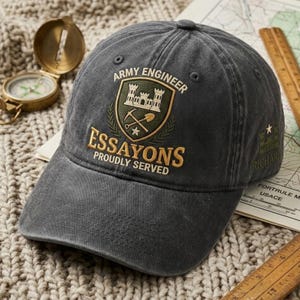 May include: A charcoal gray baseball cap featuring an embroidered Army Engineer crest with a castle, pickaxe, and shovel. The text reads ARMY ENGINEER, ESSAYONS, PROUDLY SERVED, and RICHARD. The cap has a structured crown and a curved brim.