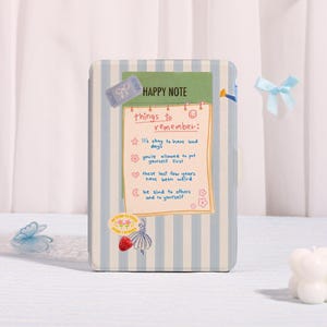 May include: A notebook with a blue and white striped cover. The notebook has a green rectangle with the words "HAPPY NOTE" and a list of reminders. Decorative stickers include a bow, flowers, and a ticket. The notebook is closed.