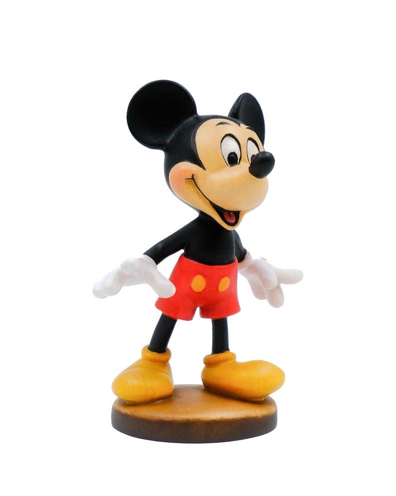 May include: A figurine of Mickey Mouse, a classic cartoon character. Mickey is depicted with black ears, a black body, red shorts, yellow shoes, and white gloves. The figurine is standing on a brown base.
