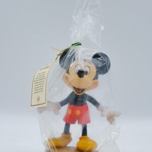 May include: A Mickey Mouse figurine in a clear plastic bag. The figurine has black ears, a yellow face, red shorts, and yellow shoes. It is standing on a brown base. A tag is attached to the bag.