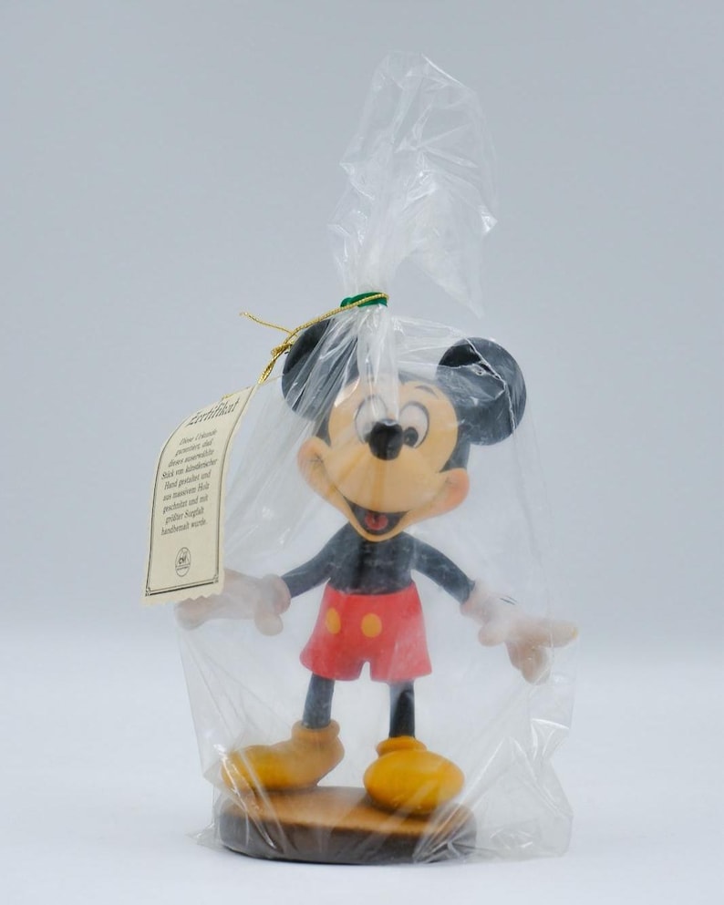 May include: A Mickey Mouse figurine in a clear plastic bag. The figurine has black ears, a yellow face, red shorts, and yellow shoes. It is standing on a brown base. A tag is attached to the bag.