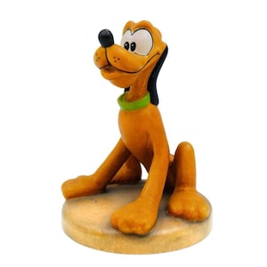 Può includere: Una statuetta di Pluto, il cane dei cartoni animati Disney, in posizione seduta. La figura è principalmente arancione con naso e orecchie neri, un collare verde e occhi bianchi. È seduto su una base circolare marrone chiaro.