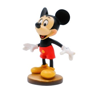 May include: A figurine of Mickey Mouse, a classic Disney character. The figure has a black head and ears, a tan face, red shorts, yellow shoes, and white gloves. It stands on a brown, circular base.