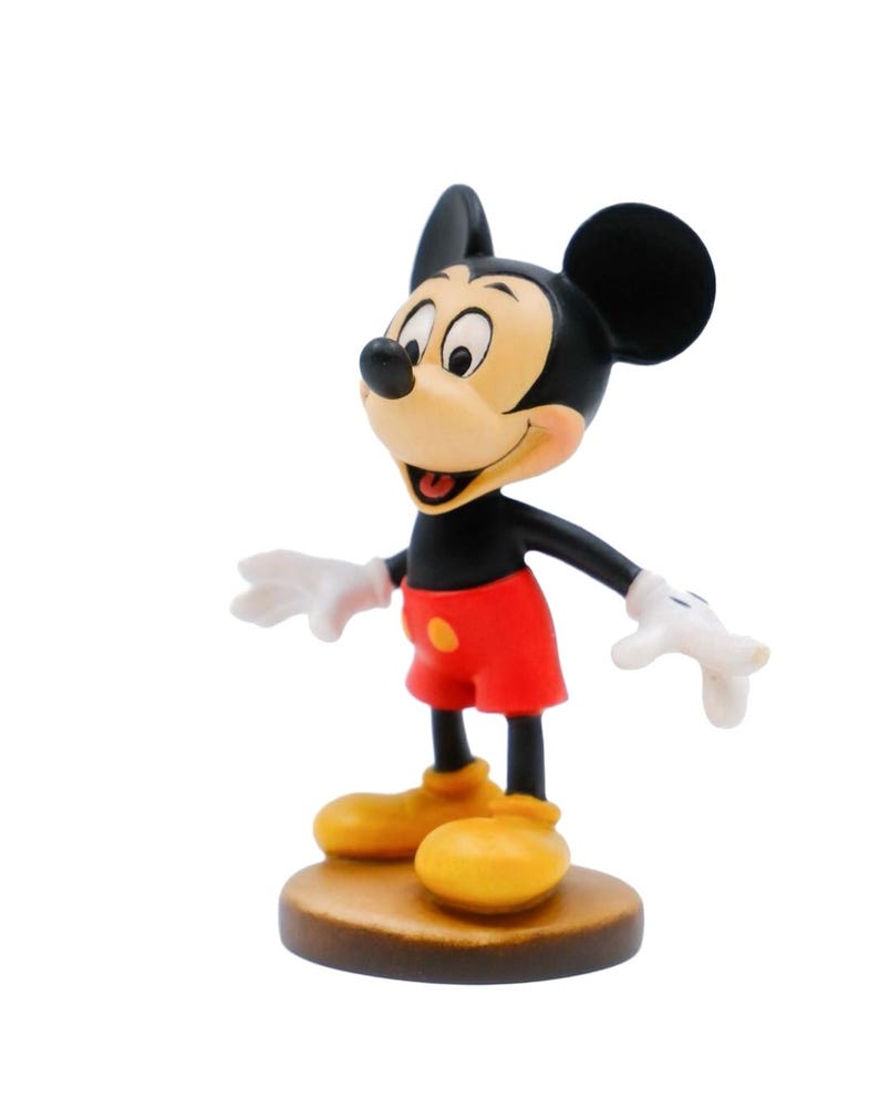 May include: A figurine of Mickey Mouse, a classic Disney character. The figure has a black head and ears, a tan face, red shorts, yellow shoes, and white gloves. It stands on a brown, circular base.
