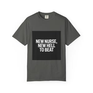 Op de afbeelding: Donkergrijs t-shirt met de witte tekst "NEW NURSE, NEW HELL TO BEAT" op een zwarte vierkant. Het t-shirt heeft korte mouwen en een ronde hals.