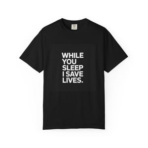 Puede incluir: Camiseta negra con el texto blanco "WHILE YOU SLEEP I SAVE LIVES." La camiseta de manga corta tiene cuello redondo. El texto está centrado en la parte delantera de la prenda.