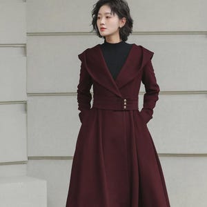 Può includere: Un cappotto lungo bordeaux con ampio colletto e cintura in vita. Il cappotto ha una gonna svasata e maniche lunghe. La modella indossa un dolcevita nero e stivali marroni. Il cappotto ha tre bottoni dorati sulla cintura.